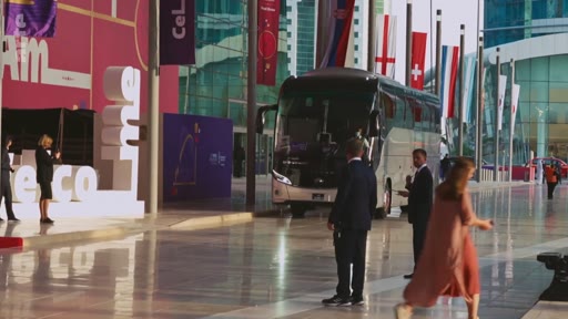 Un bus arrive devant un bâtiment moderne décoré de nombreux drapeaux nationaux, avec des personnes en costume et une femme marchant au premier plan. La scène semble se dérouler lors d'un événement international ou diplomatique.