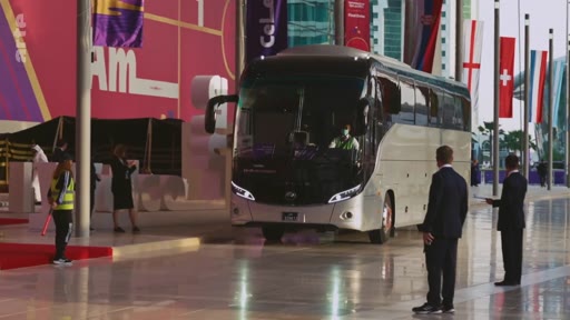 Un bus de transport officiel arrive devant un bâtiment décoré de drapeaux nationaux, probablement lors d'un événement sportif international comme la Coupe du Monde de la FIFA au Qatar. Des agents de sécurité en costume et des membres du personnel en gilet réfléchissant sont présents sur le parvis.