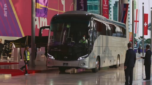 Un bus de transport arrive devant un bâtiment décoré aux couleurs de la Coupe du Monde 2022 au Qatar, avec des drapeaux nationaux visibles en arrière-plan.