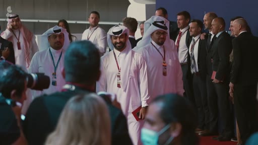 Des officiels qataris en tenue traditionnelle, dont certains portent des badges d'accréditation, marchent sur un tapis rouge lors d'un événement officiel, entourés de photographes et d'autres personnalités en costume.