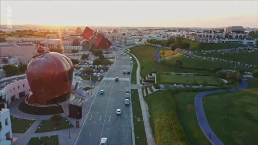 Vue aérienne en plongée du Katara Cultural Village à Doha, au Qatar, mettant en évidence l'architecture distinctive du site, notamment le bâtiment en forme de dôme cuivré et les espaces paysagers au coucher du soleil.