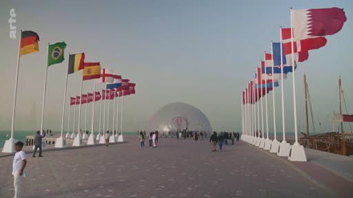 Vue d'ensemble d'une esplanade au Qatar bordée de drapeaux de différentes nations, menant à une structure architecturale en forme de dôme, dans le contexte de la Coupe du Monde de la FIFA 2022.