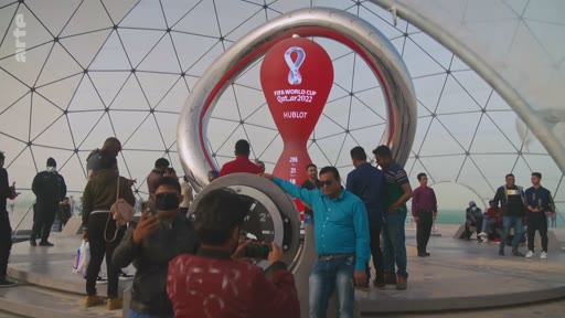 Des touristes et des visiteurs se rassemblent devant l'horloge compte à rebours officielle de la Coupe du Monde de la FIFA Qatar 2022, parrainée par Hublot, située sous une structure en dôme à Doha. L'image montre l'effervescence autour de cet événement sportif mondial majeur.