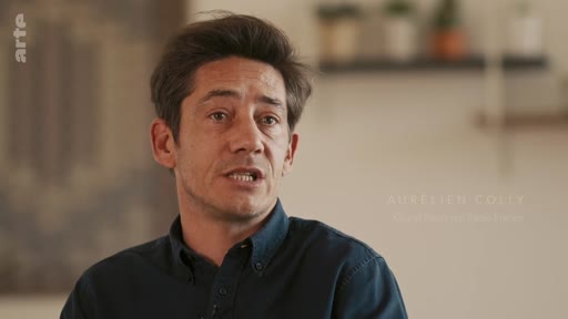 Aurélien Folly est interviewé dans une séquence filmée, portant une chemise sombre. Il s'exprime face caméra dans un cadre intérieur.