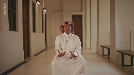 Un homme portant une tenue traditionnelle arabe et un keffieh est assis dans un couloir aux murs blancs pour une interview. Il s'exprime avec les mains dans un cadre formel.