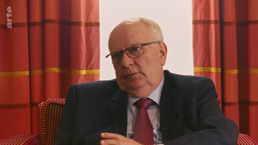 Mikhail Gorbatchev, ancien dirigeant de l'Union soviétique, est interviewé dans un cadre formel. Il porte un costume sombre et une cravate rouge, s'exprimant face caméra.