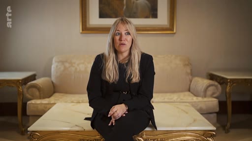 Une femme blonde, vêtue d'une veste noire, est assise sur une table basse dans un salon au décor classique, lors d'une interview pour la chaîne Arte.