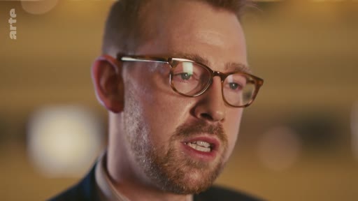 Un homme d'âge moyen, portant des lunettes et une barbe légère, est filmé en gros plan lors d'une interview télévisée. Il s'exprime face caméra dans un cadre formel.