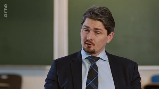 Un homme en costume sombre et cravate à motifs s'exprime face caméra lors d'une interview télévisée, probablement dans un cadre scolaire ou académique avec un tableau vert en arrière-plan.