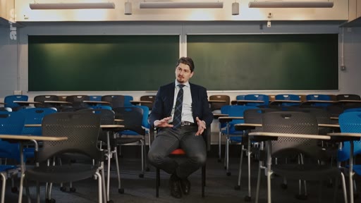 Un homme en costume et cravate est assis sur une chaise au milieu d'une salle de classe vide, devant des tableaux noirs, lors d'une interview.