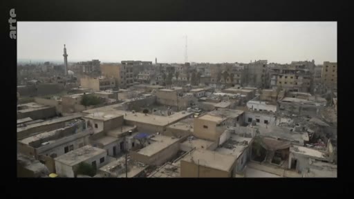 Vue panoramique en plongée sur un quartier urbain dense au Moyen-Orient, marqué par des traces de destruction et des bâtiments dégradés, avec un minaret visible à l'arrière-plan.
