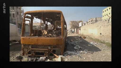 Un bus totalement détruit et calciné est stationné dans une rue dévastée en Syrie, témoignant des conséquences de la guerre civile syrienne.