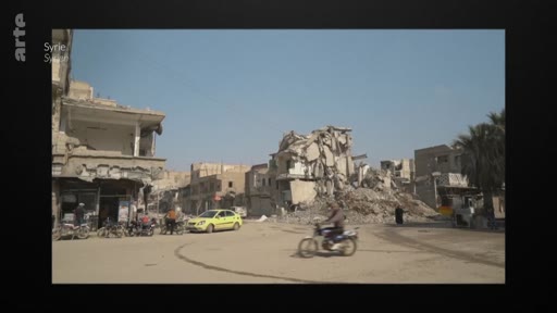Vue d'une rue en Syrie montrant des bâtiments détruits et des décombres, avec un taxi jaune et un motocycliste circulant au milieu des ruines. Cette image illustre les conséquences dévastatrices du conflit syrien sur l'infrastructure urbaine.