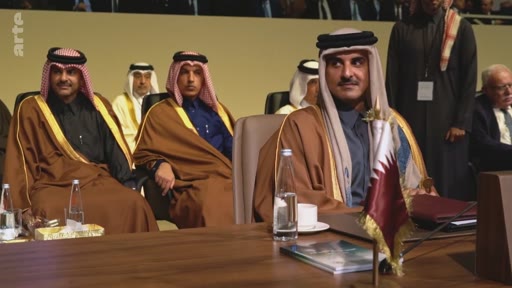 L'émir du Qatar, Tamim ben Hamad Al Thani, est assis à une table lors d'une conférence ou d'un sommet officiel, entouré d'autres dignitaires portant des tenues traditionnelles arabes. Le drapeau du Qatar est visible sur la table devant lui.