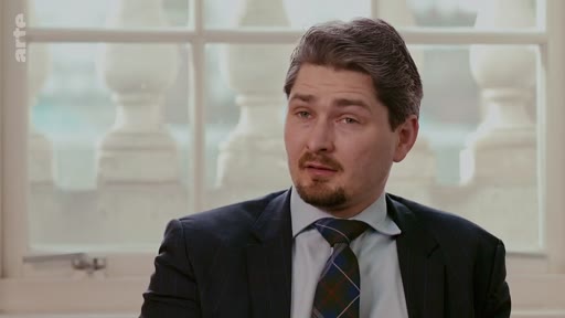 Un homme en costume sombre et cravate à carreaux est interviewé dans un cadre formel, probablement un bureau ou un studio, avec une fenêtre en arrière-plan.