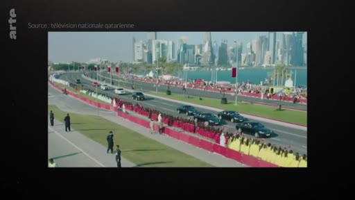 Un cortège de voitures officielles circule sur une avenue de Doha, au Qatar, avec la skyline de la ville en arrière-plan. Des personnes en tenue traditionnelle sont alignées le long de la route, probablement lors d'un événement officiel ou d'une célébration nationale.