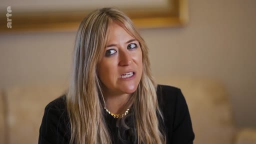 Une femme blonde aux cheveux longs s'exprime face caméra lors d'une interview télévisée, portant un haut noir et un collier doré.