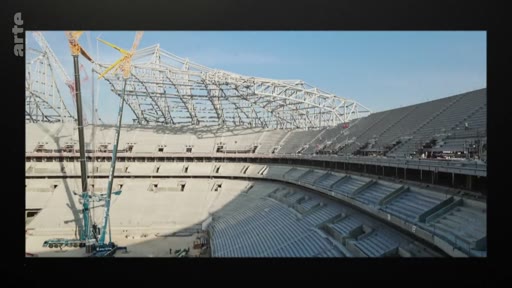 Vue en plongée d'un grand stade de football en cours de construction, montrant la structure métallique de la toiture et les gradins en béton, avec une grue de chantier visible sur le côté.