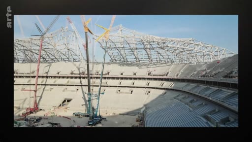 Vue panoramique d'un grand stade de football en cours de construction, montrant la structure métallique du toit et plusieurs grues de chantier sur le terrain.