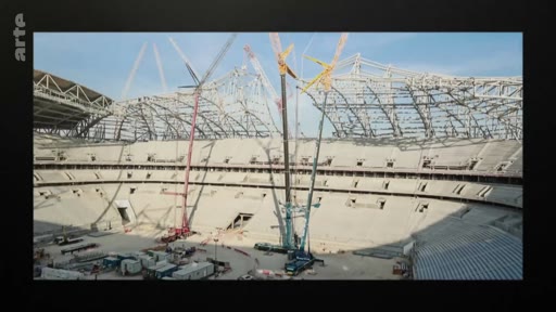 Vue d'ensemble d'un grand stade de football en cours de construction, montrant la structure métallique de la toiture et plusieurs grues de chantier sur le terrain. Ce chantier illustre les travaux d'infrastructure sportive à grande échelle.