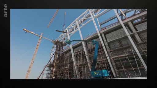 Vue en contre-plongée d'un chantier de construction d'un grand bâtiment, montrant des grues et une nacelle élévatrice en action sous un ciel bleu dégagé.