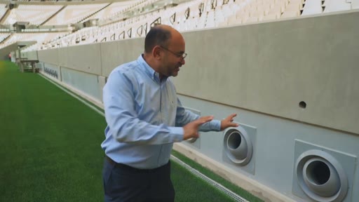 Un homme en chemise bleue présente le système de ventilation intégré aux murs d'un stade de football moderne. Il explique le fonctionnement des bouches d'aération situées au niveau de la pelouse.