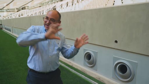 Un homme chauve portant des lunettes et une chemise bleue explique le fonctionnement du système de ventilation intégré aux tribunes d'un stade de football moderne.