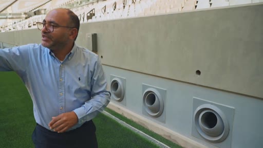 Un homme portant des lunettes et une chemise bleue visite les abords d'un terrain de football, montrant les systèmes de ventilation intégrés au mur du stade.
