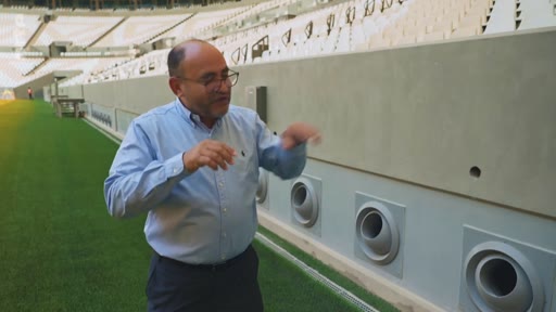 Un homme, portant des lunettes et une chemise bleue, gesticule en parlant devant les tribunes d'un stade de football moderne. Il se trouve sur la pelouse, près d'un mur équipé de bouches d'aération.