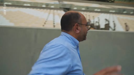 Un homme chauve portant des lunettes et une chemise bleue marche devant les gradins d'un stade, probablement lors d'un reportage sur des infrastructures sportives.