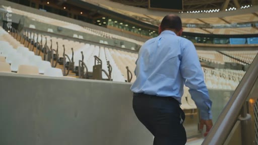 Un homme en chemise bleue et pantalon sombre monte les escaliers dans les tribunes d'un grand stade de sport vide. La vue montre l'architecture intérieure de l'enceinte sportive.
