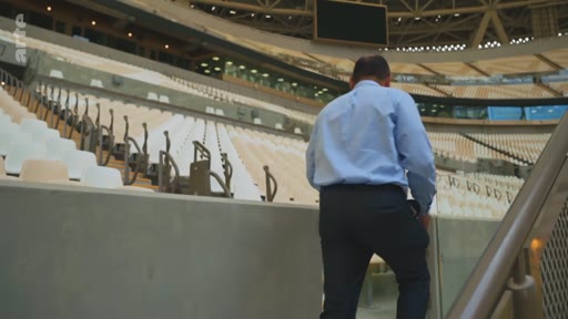 Un homme en chemise bleue et pantalon sombre monte les escaliers dans les tribunes d'un stade de football moderne et vide. L'architecture du stade est visible en arrière-plan avec des sièges blancs et beiges.