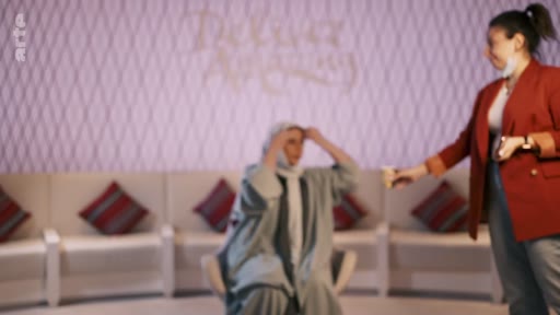 Une femme portant un voile et une tenue traditionnelle est assise sur un plateau de télévision, ajustant sa coiffe, tandis qu'une autre femme en veste rouge se tient debout à ses côtés. La scène se déroule dans un studio avec un décor épuré et des canapés en arrière-plan.