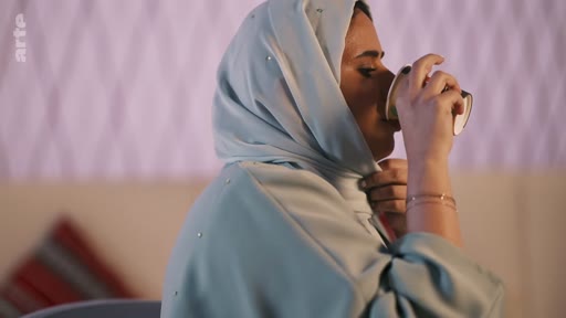 Une femme portant un hijab de couleur claire boit dans un gobelet en papier lors d'une séquence filmée. Elle porte des bijoux discrets et est filmée en plan rapproché.