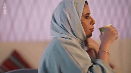 Une femme portant un hijab de couleur claire est filmée de profil alors qu'elle boit dans un gobelet en carton. La scène se déroule dans un intérieur avec un éclairage tamisé.