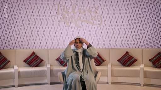Une femme portant une abaya et un voile est assise sur un plateau de télévision, devant un mur décoré du logo 'Deluxe Arabia'. Elle ajuste son voile tout en étant filmée dans un studio au décor épuré avec des coussins à motifs traditionnels.