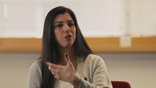 Une femme aux cheveux longs bruns s'exprime en faisant un geste de la main lors d'une interview filmée en intérieur.