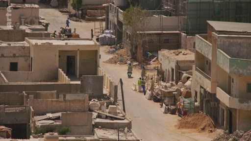 Vue en plongée d'une rue étroite dans un quartier en développement avec des maisons en construction, des ouvriers et des habitants circulant à pied ou en deux-roues.