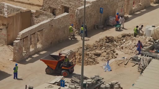 Vue aérienne d'un chantier de construction ou de rénovation urbaine où des ouvriers en gilets de sécurité manipulent des matériaux et utilisent un petit tombereau motorisé près de murs en pierre ancienne.