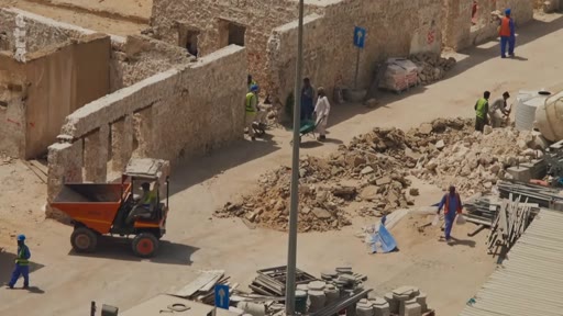 Vue aérienne d'un chantier de construction où des ouvriers s'activent avec des brouettes et un petit engin de chantier orange, dans un environnement urbain en cours de rénovation ou de démolition.