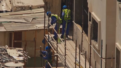 Des ouvriers du bâtiment portant des casques bleus et des gilets de sécurité travaillent sur un échafaudage métallique le long d'un bâtiment en rénovation.