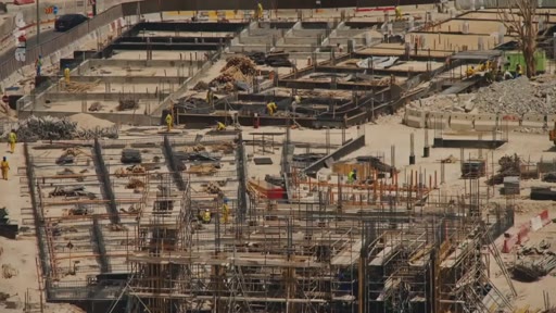 Vue aérienne d'un vaste chantier de construction montrant les fondations et les structures en béton armé en cours d'assemblage par des ouvriers. Le site présente plusieurs zones de travaux avec des armatures métalliques et des coffrages.