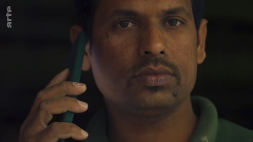 Un homme d'origine indienne, portant un polo vert, est filmé en gros plan alors qu'il communique au téléphone. L'image est sombre et met l'accent sur son expression sérieuse.