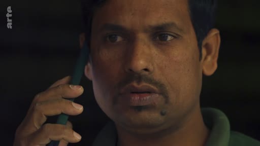 Un homme d'origine indienne est filmé en gros plan alors qu'il est en pleine conversation téléphonique, affichant une expression sérieuse et préoccupée.