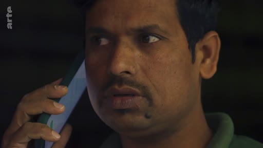 Un homme d'origine indienne est filmé en gros plan alors qu'il est en pleine conversation téléphonique, exprimant une certaine préoccupation.
