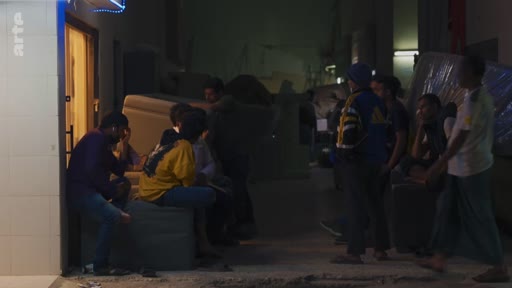 Un groupe de personnes, dont certaines sont assises sur des meubles, se réunit devant une entrée éclairée dans une rue sombre. La scène suggère une situation de précarité ou un rassemblement informel en extérieur.