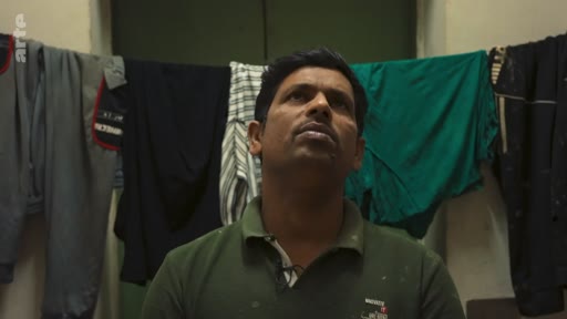 Un homme d'origine indienne est filmé en plan moyen, regardant vers le haut, dans un intérieur modeste où des vêtements sont suspendus pour sécher en arrière-plan. Cette séquence semble issue d'un reportage sur les conditions de vie ou le travail des migrants.