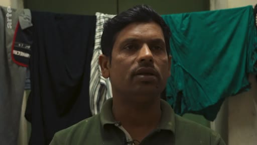 Un homme d'origine indienne s'exprime face caméra dans ce qui semble être un logement modeste, avec du linge séchant en arrière-plan. Il s'agit d'un portrait réalisé dans le cadre d'un reportage télévisé.