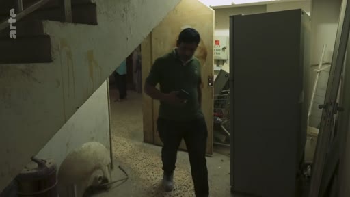 Un homme marche dans un espace intérieur sombre et encombré, possiblement un sous-sol ou un bâtiment en ruine, filmé par une équipe de reportage d'Arte.