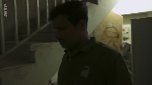 Un homme non identifié se déplace dans un escalier intérieur sombre, filmé dans un style documentaire. L'image présente une atmosphère austère et utilitaire, typique d'un reportage sur le terrain.